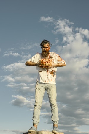 Halloween hipster with pumpkin standing high on grey sky. Zombie man in torn clothes with red blood. War soldier or victim. Horror and violence concept. Trick or treat.の写真素材