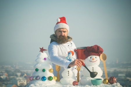 Sad man in santa hat ladling soup on winter day. Hipster with ladle in white uniform. Cooking and diet concept. Cook, snowmen and snow xmas tree on blue sky. Christmas and new year holidays food.の写真素材