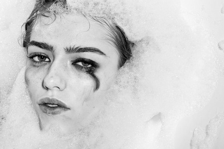 Closeup view portrait of one beautiful sensual sexy attractive young woman with wet bright makeup lying in bath tub with water and lots of white soap foam looking away, horizontal pictureの写真素材