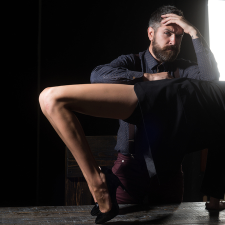Guy at table with female legs. Love and relations, dominating. Legs of woman in shoes at man with beard. Romance and couple in love. Man in shirt isolated on black background.の写真素材