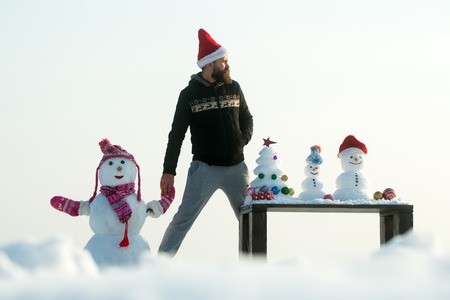 Winter fun and holidays celebration concept. Hipster in red santa hat. Snow sculptures and xmas tree on wooden table. Christmas and new year. Man and snowman holding hands on white sky.の写真素材