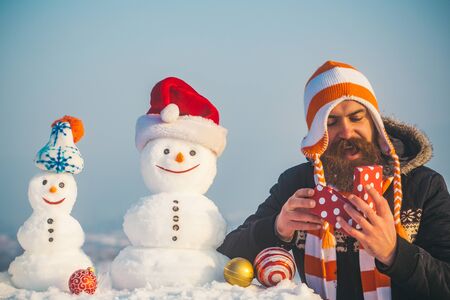 Surprised hipster in hat holding present box. Christmas and new year. Snow sculptures on blue sky. Winter holidays celebration concept. Man and snowmen in santa cap.の写真素材