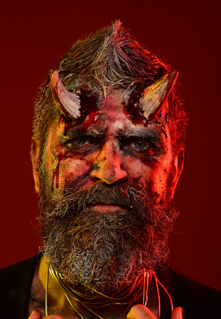 Halloween man devil on red background. Satan with beard, blood, wounds on face. Demon with bloody horns on head. Hell, death, evil, horror concept. Holiday celebration, cosplay.の写真素材