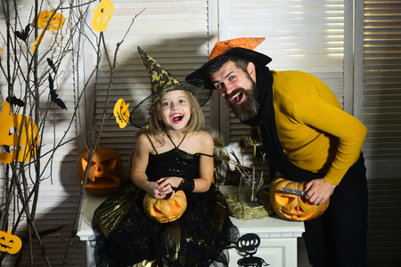 Father and daughter with Halloween decor. Wizard and little witch in hats hold jack o lantern. Girl and bearded man with cheerful faces on red spooky carnival room background. Halloween party conceptの写真素材