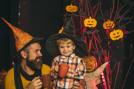 Halloween party and celebration. Holiday and celebration. Boy kid with happy face and bearded man at pumpkin. Halloween man and child in witch hat. Father and small son drink from tea or milk cup.の写真素材