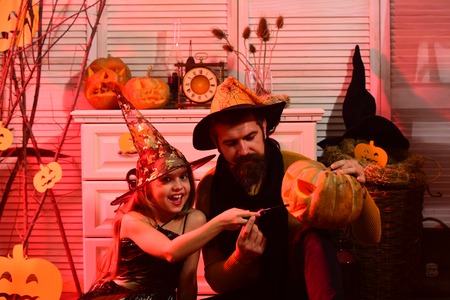 Wizard and little witch in black hats carve pumpkin. Girl and bearded man with happy faces on red spooky carnival room background. Father and daughter with Halloween decor. Halloween party conceptの写真素材