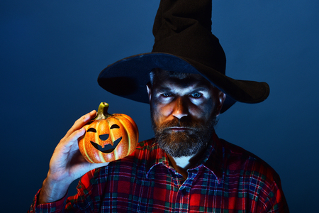 Halloween man in witch hat holding pumpkin on blue background. Wizard hipster with beard and jack o lantern. Autumn holiday celebration. Trick or treat. Mystery and magic concept.の写真素材