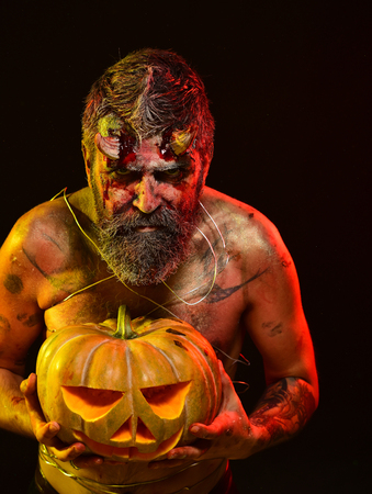 Halloween satan stare with bloody horns, beard, red blood, wounds. Man demon hold pumpkin on black background. Trick or treat. Darkness and light concept. Devil with jack o lantern.の写真素材