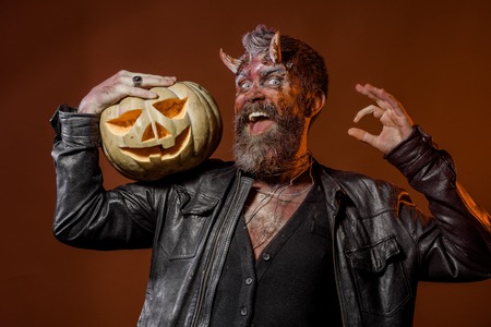 Halloween devil with bloody horns show ok hand gesture. Happy man demon hold pumpkin on brown background. Autumn holidays concept. Satan with beard, wounds, red blood on face. Trick or treat.の写真素材