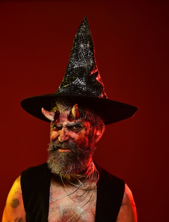 Halloween devil hipster with beard, blood, wounds on face. Man with satan horns in witch hat on red background. Hell, death, evil, horror concept. Might, magic, witchcraftの写真素材