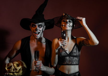 Halloween couple with makeup on brown background. Couple in love with pumpkin and wine glass. woman and man has happy face in witch hat. Halloween Holiday and celebration. Zombie party and horror.の写真素材