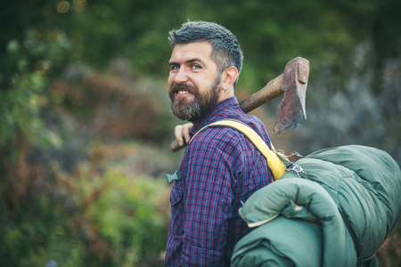 Man lumberjack happy smiling with axe and backpack on summer day on natural landscape, bokeh effect. Logging and chopping concept, copy spaceの写真素材