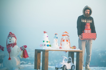 Unhappy man holding gift boxes. Snowman with shopping bags on blue sky. Christmas and new year presents concept. Snowy sculptures and xmas tree on wooden table. Toy car on snow.の写真素材