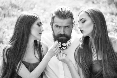 handsome man in white shirt with dandelion flowers in beard with two young pretty women in violet dresses on green grass sunny day outdoorの写真素材