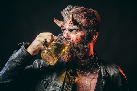 Halloween hipster with satan horns hold glass mug. Devil with red blood and wounds on face. Holiday celebration and party. Bad habits and addiction concept. Man demon drink beer on black background.の写真素材