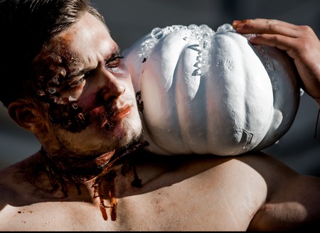 Halloween Party and bloody horror. demon with muscular body and makeup. man with zombie face sunny outdoor. Holiday and celebration. Halloween guy with white traditional pumpkin.の写真素材
