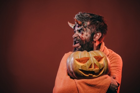 Halloween satan show tongue with horns, beard, blood, wounds. Man devil hold pumpkin on brown background. Darkness and light concept. Demon in blanket with jack o lantern. Trick or treat, copy spaceの写真素材