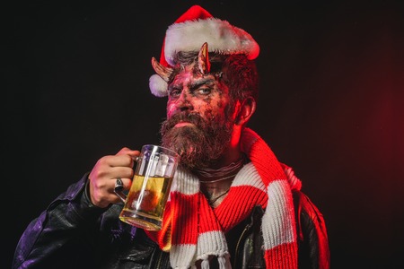 Halloween christmas holiday, party celebration. Hipster with satan horns hold glass mug. Devil in santa hat and scarf. Man demon drink beer on black background. Bad habits and addiction concept.の写真素材