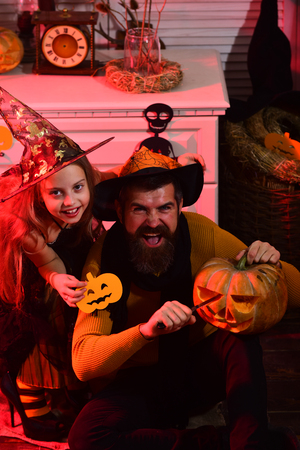 Girl and bearded man with happy faces on red spooky carnival room background. Halloween party concept. Dad and kid in witch hats hold carved pumpkins. Wizard and little witch with Halloween decorの写真素材