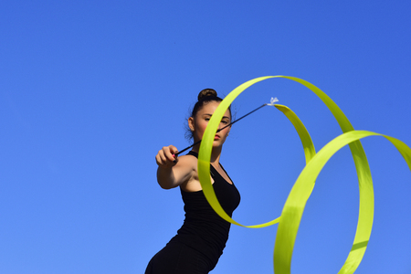 Woman gymnast in black sportswear with green ribbon. Sport and success. Workout of girl on blue sky background. Gymnastic school and energy. Flexibility in acrobatics and fitness health.の写真素材