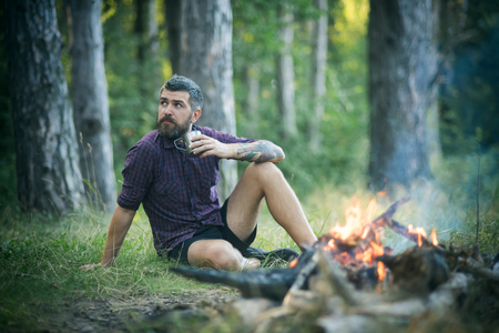 Hipster hiker with mug relax at bonfire in forest. Man traveler drink tea at campfire flame. Summer vacation concept. Camping, hiking, lifestyle. Travel, traveling, wanderlust.の写真素材