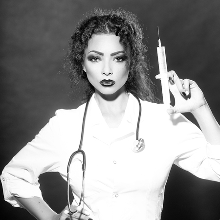 Closeup view portrait of one serious sexual beautiful brunette medical worker with stethoscope in white nurse uniform holding injecting suringe or drugs in studio, square pictureの写真素材