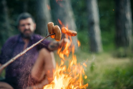 Sausages roasted on bonfire flame with blurred man hipster on natural background. Picnic, barbecue, cooking food concept. Summer camping, hiking, vacation.の写真素材