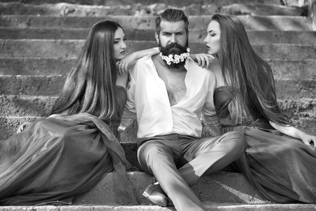 handsome man in white shirt with dandelion flowers in beard with two young pretty women in violet dresses on stony stairs sunny day outdoorの写真素材