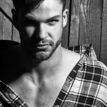 One handsome serious young sensual sexy man in checkered shirt with beard and bare chest looking forward in studio on wooden wall backdrop, square pictureの写真素材