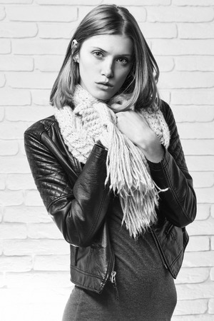 young pretty woman or fashionable sexy girl with cute face and blonde hair in scarf and leather jacket on white brick wall backgroundの写真素材