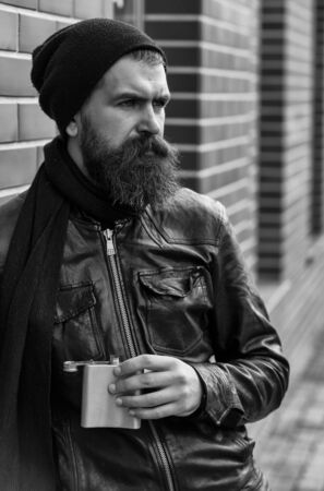Bearded man, long beard, brutal caucasian hipster with moustache holding metallic flask, jar, has serious face, in black leather jacket, hat, scarf on brick wall background outdoorの写真素材
