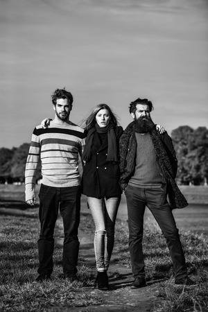 Young people friends of pretty cute sexy girl or woman and two bearded men outdoors in park on autumn sunny day on natural blue sky backgroundの写真素材