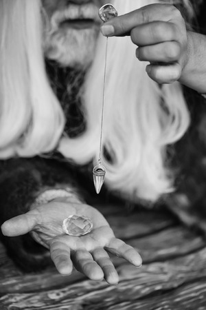 Old bearded man wizard with long white hair and beard holding blue gem stone and silver pendant for hypnosis on wooden backgroundの写真素材