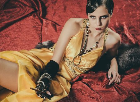retro look and pinup. Girl in yellow dress and fur with mouthpiece. Woman with stylish retro hair, makeup and cigaterre holder. Pin up pretty fashion model on red blanket. Beauty and vintage fashion.の写真素材
