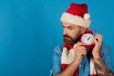 Christmas new year, xmas holidays celebration. Hipster in santa hat, scarf on blue background. Time to celebrate concept. Man hold alarm clock. Countdown to midnight, copy spaceの写真素材
