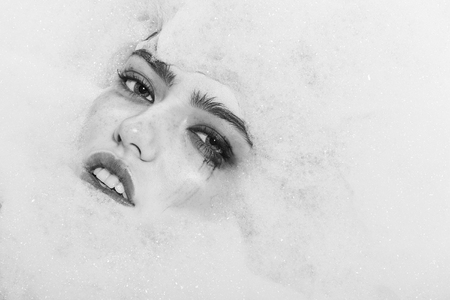 Closeup view portrait of one beautiful sensual sexy attractive young woman with wet bright makeup lying in bath tub with water and lots of white soap foam looking away copy space, horizontal pictureの写真素材