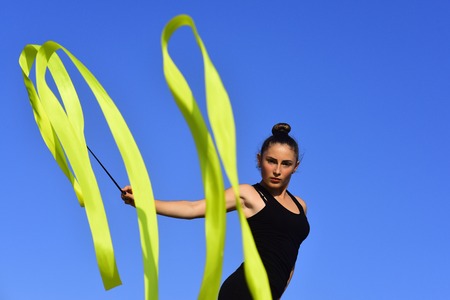 Flexibility in acrobatics and fitness health. Workout of girl on blue sky background. Gymnastic school and energy. Sport and success. Woman gymnast in black sportswear with green ribbon.の写真素材