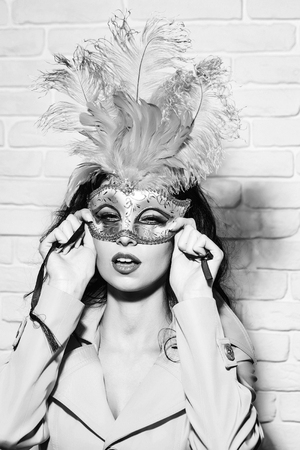 Pretty girl or cute sexy woman has brunette hair in golden festive carnival mask with feathers and yellow coat on white brick wall backgroundの写真素材