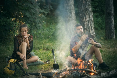 Girl or woman use mobile phone. New technology or archaism concept. Couple of hikers relax at bonfire in forest. Man hipster sharpen axe. Camping, hiking, summer vacation.の写真素材