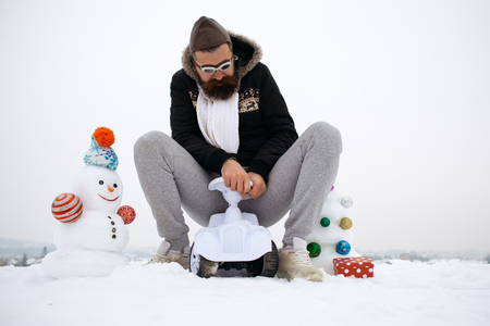 Hipster riding toy car on white sky. Bearded man in pilot glasses and hat. Snowman and snow xmas tree on winter landscape. Holiday celebration concept. Christmas and new year fun.の写真素材