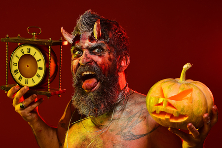 Halloween autumn holidays concept. Man demon hold pumpkin and clock. Satan show tongue with beard, wounds, blood, tattoo on chest. Devil with bloody horns on red background. Time to celebrate.の写真素材