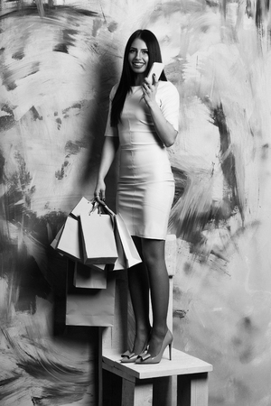 pretty happy girl or cute fashionable woman with long brunette hair in sexy pink dress with shopping bags or packages holds mobile or cell phone on chair on colorful wall backgroundの写真素材