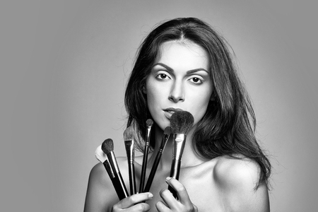 young woman with pretty face and long brunette hair with sexy bare shoulders of female body holding fashionable makeup brush set in studio on grey background, copy spaceの写真素材