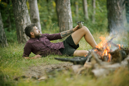 Camping, hiking, lifestyle. Hipster hiker with mug relax at bonfire in forest. Summer vacation concept. Travel, traveling, wanderlust. Man traveler drink tea at campfire flame.の写真素材