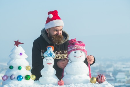 Winter holidays celebration concept. Santa man putting hat on snowman. Christmas and new year decorations. Snow xmas tree and sculptures on blue sky. Hipster in red santa claus cap.の写真素材