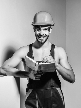 Happy man smiling handsome builder repairman craftsman foreman or construction worker in orange hard hat and overall keeps accounting book on grey backgroundの写真素材