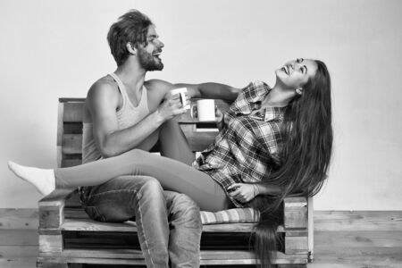 Young happy couple of pretty girl or cute woman, with brunette long hair and handsome man or muscular macho with muscular torso, sits on wooden with tea cups, pallet sofa on grey backgroundの写真素材