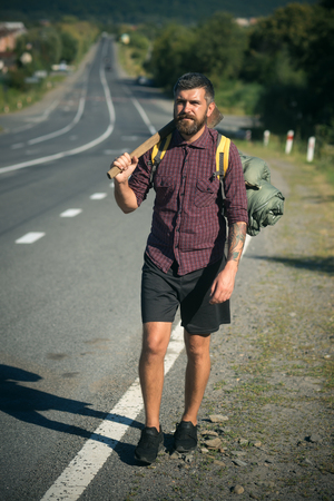 Hipster man with axe and tourist backpack stand on road side on sunny day. Summer vacation, active lifestyle and traveling conceptの写真素材