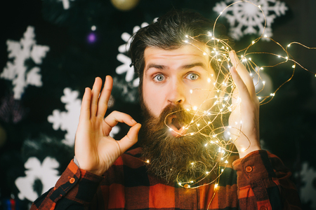Party and celebration. Garland on santa claus man as decoration show ok gesture. Winter holiday and xmas. New year guy with illuminated wire. Christmas man with beard on surprised face and garland.の写真素材