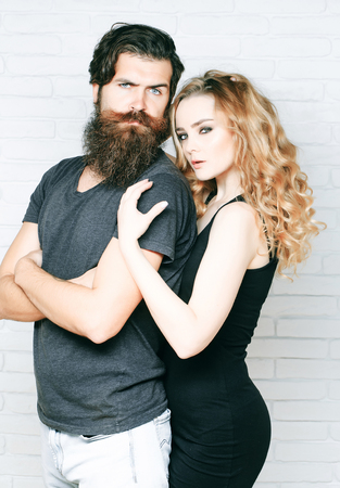 Hipsterism, subculture, trend. Man with beard and woman with long blond hair. Fashion, beauty, style concept. Girl and bearded hipster. Couple in love hug on white brick wall.の写真素材
